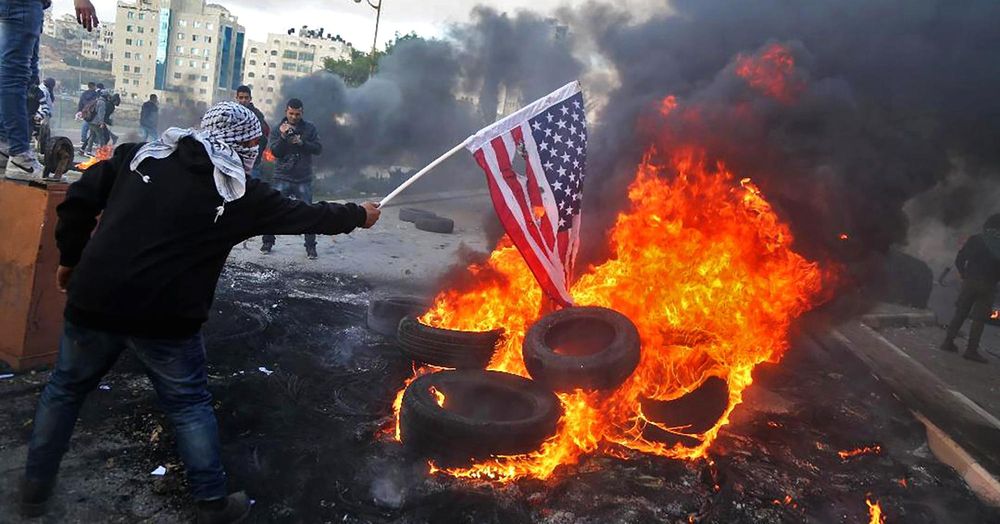 A person in a black jacket and keffiyeh waves an American flag near burning tires emitting thick black smoke, with other individuals in the background amid urban unrest.