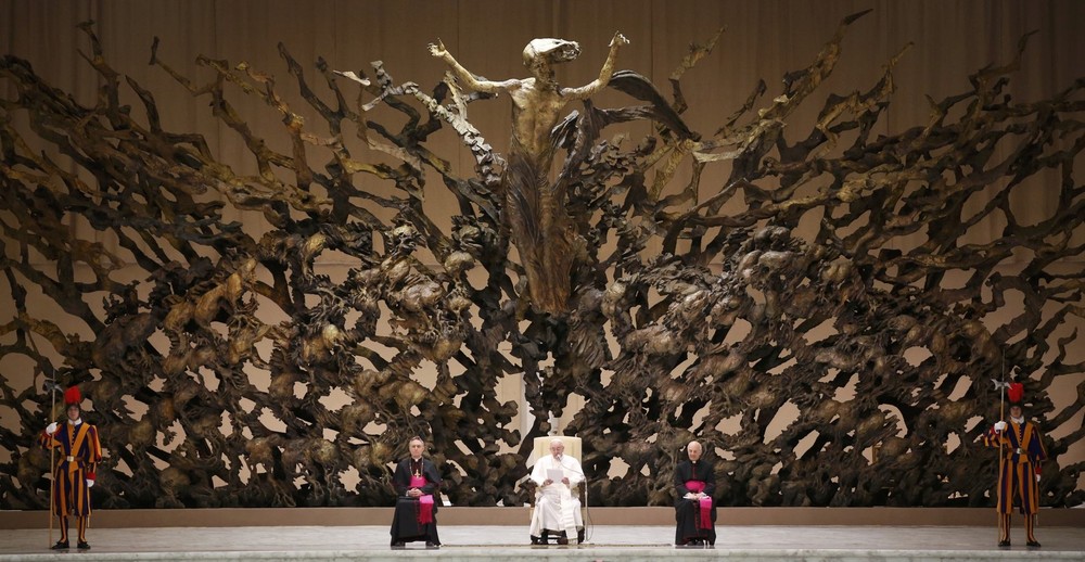 A large, ornate sculpture resembling a tree with extensive branches fills the background. Several figures dressed in various ceremonial attires, including two in colorful uniforms resembling Swiss guards, two clerics seated with one in white and the other in purple, and one woman in black using a wheelchair, are positioned in front of the sculpture.