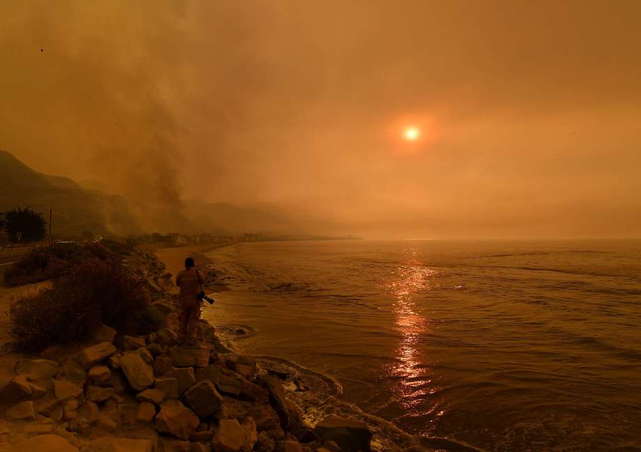A person stands on a rocky shoreline, watching a dramatically hued sky due to smoke, with the sun appearing as a faint, reddish orb. Smoke obscures the mountains in the background, and the ocean reflects the muted sunlight creating a surreal, amber-tinted environment.