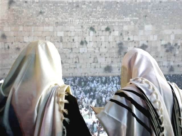 Two individuals in traditional Jewish prayer shawls with their backs to the camera overlook a large gathering of people at the Western Wall in Jerusalem.