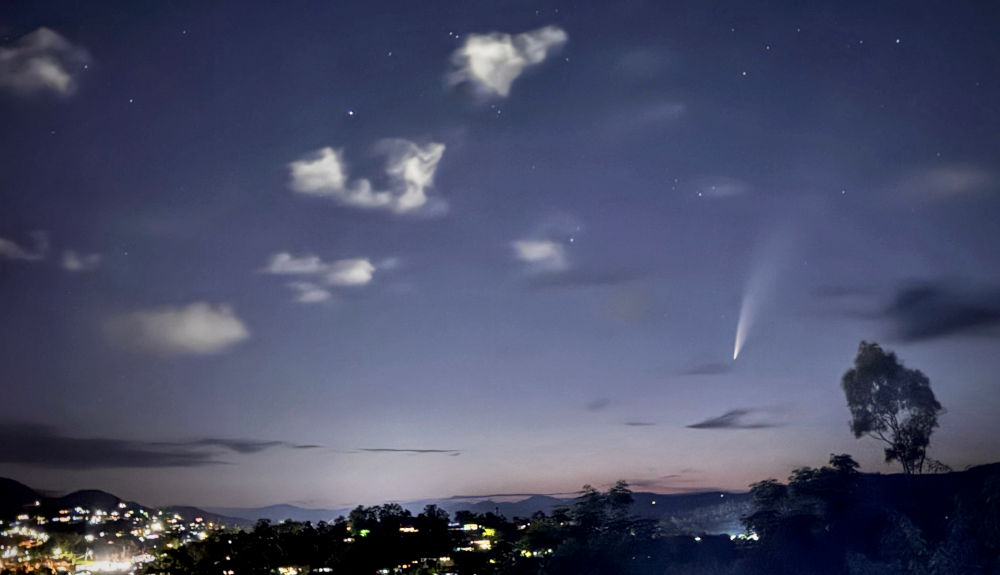 Nighttime landscape showing a luminous comet with a long tail visible in the dark sky, above softly lit hillside town. Scattered clouds and stars are also visible, with a silhouette of a solitary tree on the right.