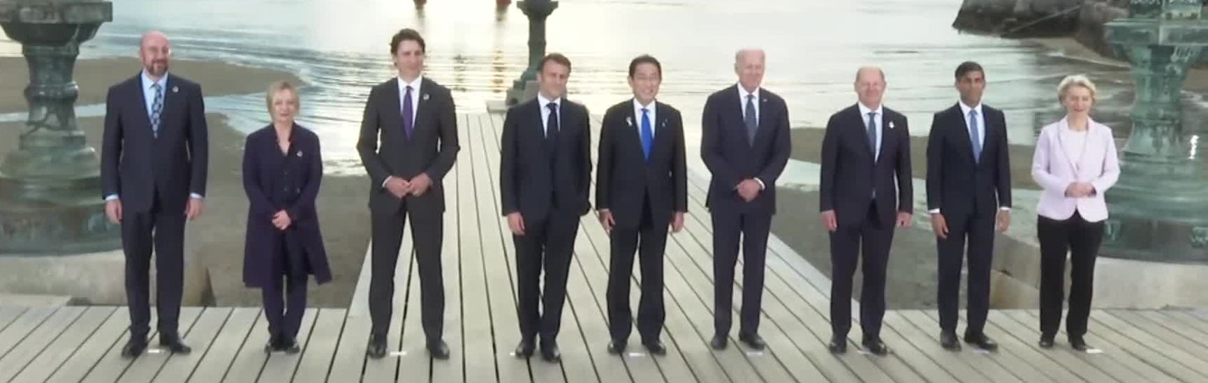 A group of nine diverse leaders, four women and five men, stands in a line on a wooden pier, dressed in formal attire. They are posed against a soft backdrop with a reflecting water body under cloudy skies.
