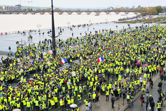 A large crowd of people wearing bright yellow vests gathered by a broad river, with several bridges in the background under an overcast sky. Some individuals are holding flags.