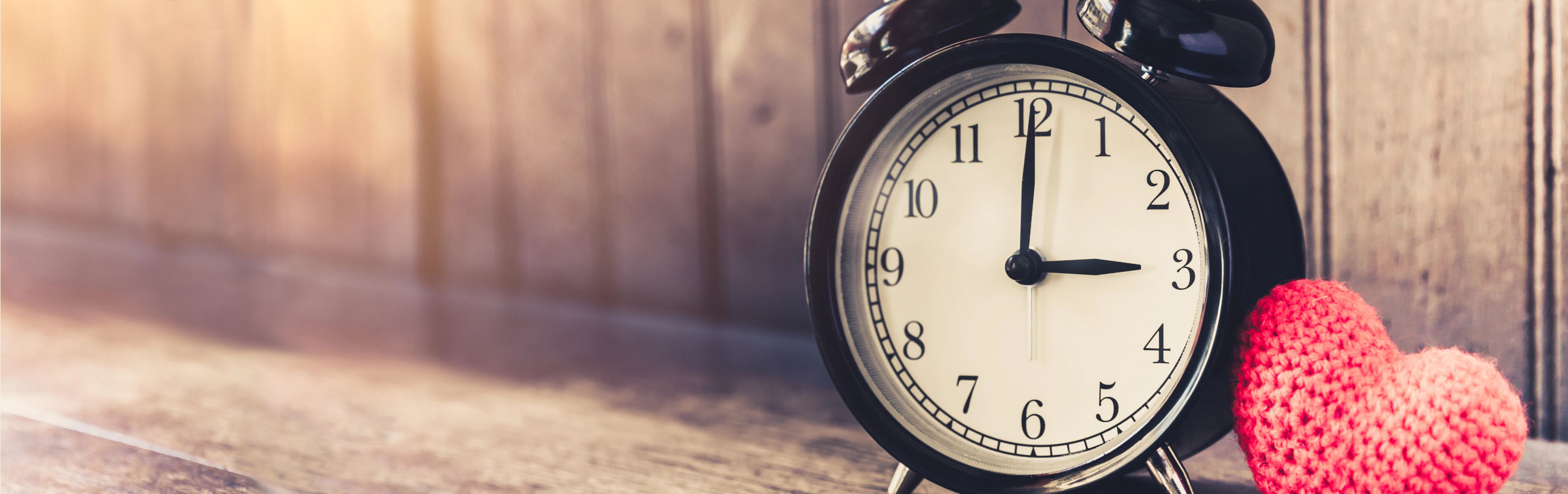 A traditional black analog clock showing ten minutes past nine, positioned next to a small, handmade red heart placed on a wooden surface, illuminated by warm, soft light.