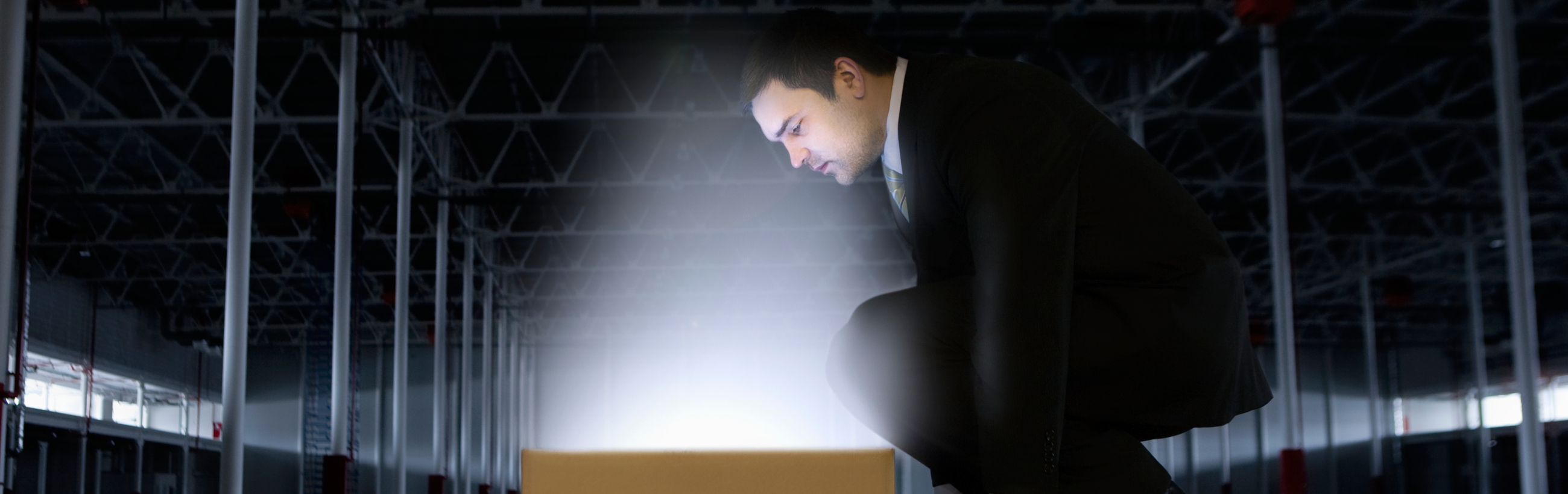 A man in a dark suit crouches beside a glowing box in a dimly lit warehouse, reminiscent of celestial phenomena akin to observing a singular radiant point in the vast expanse of a night sky.