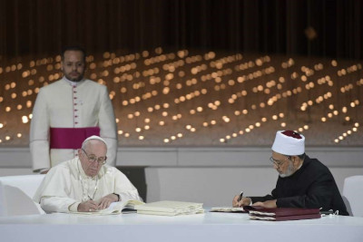 Two religious leaders, one in white robes and the other in black with a white cap, sit at a table signing documents. Behind them, a backdrop of numerous small lights provides a serene setting, reminiscent of stars in the night sky.