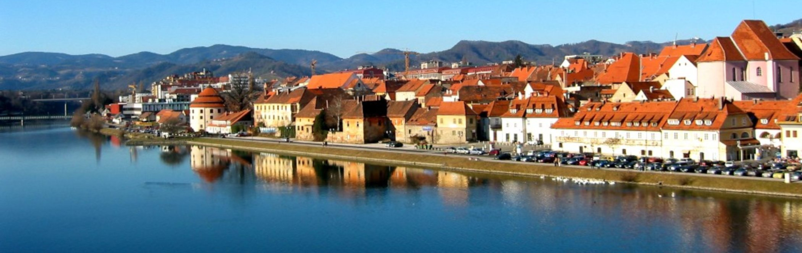 Panoramic view of a peaceful riverside town under a clear blue sky, featuring mountains in the background, with historical architecture reflecting off the calm river surface.