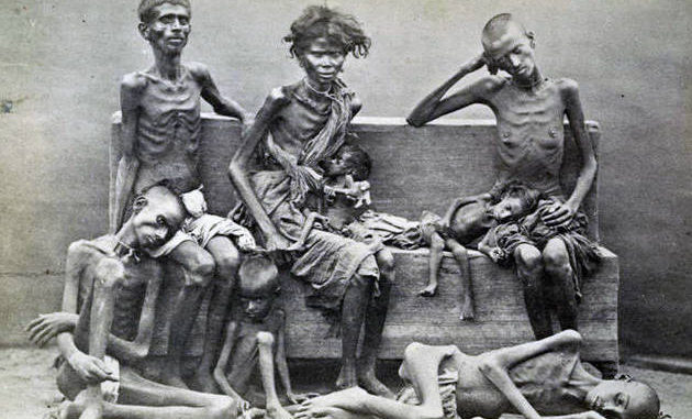 Historical photograph depicting a group of extremely emaciated men and women sitting and lying on a simple wooden bench against a plain background.