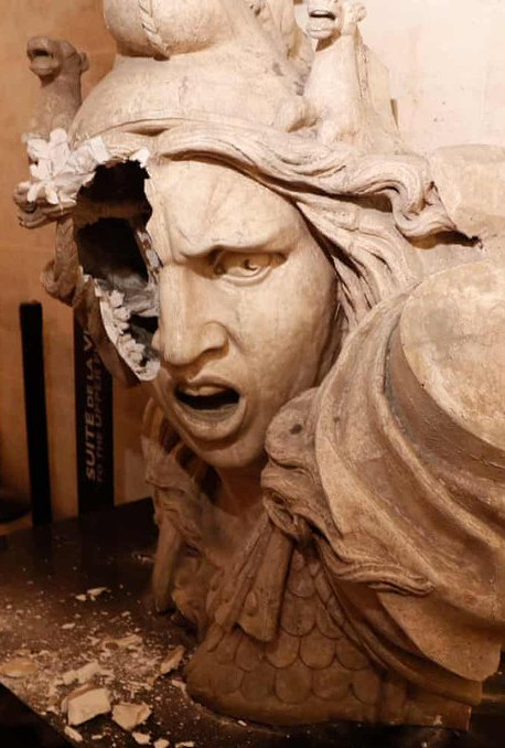 A detailed sculpture of an expressive human figure with intricate armor and celestial adornments; the piece has a visible damage, with part of the head and helmet missing, exposing the inner layers of the sculpture. The figure's facial expression is intense and fierce, suggesting a connection to a mythological or heavenly theme rooted in ancient cultural depictions.