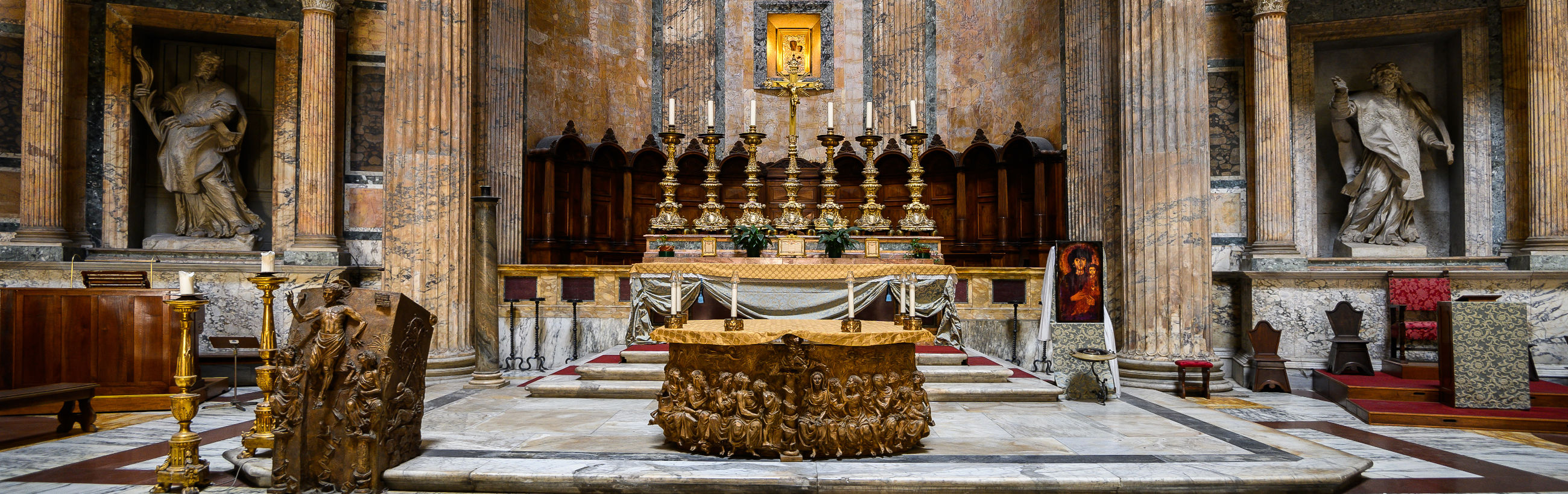 Interior view of a grand historical basilica, showcasing an ornate altar adorned with golden elements and surrounded by massive marble columns. Statues are nested within niche spaces between the columns, and religious art enhances the sanctified atmosphere.