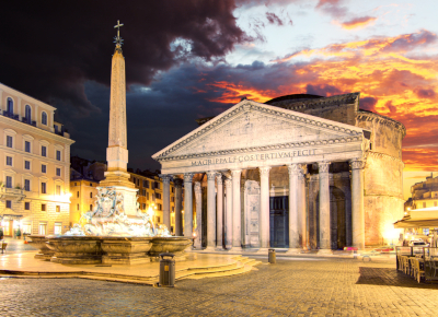 A majestic view of the Pantheon in Rome, illuminated brilliantly during twilight. This ancient temple, initially dedicated to all deities under the configuration of the celestial sphere, stands proudly with its classic Corinthian columns and triangular pediment. A towering obelisk, rooted in a sculpted fountain, rises in the foreground under a dramatically fiery sky.