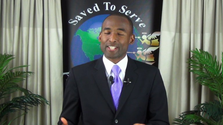 Un hombre con traje negro y corbata violeta habla con una sonrisa amable frente a una pancarta con la leyenda "Salvados para servir" que presenta un gráfico de la Tierra y varios símbolos celestiales, incluidas ilustraciones que se asemejan a las constelaciones del Mazzaroth representadas alrededor de la Tierra.