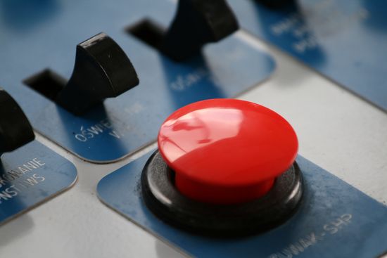 Primer plano de un botón rojo intenso en una máquina con varios botones negros etiquetados en inglés, sobre un fondo azul.