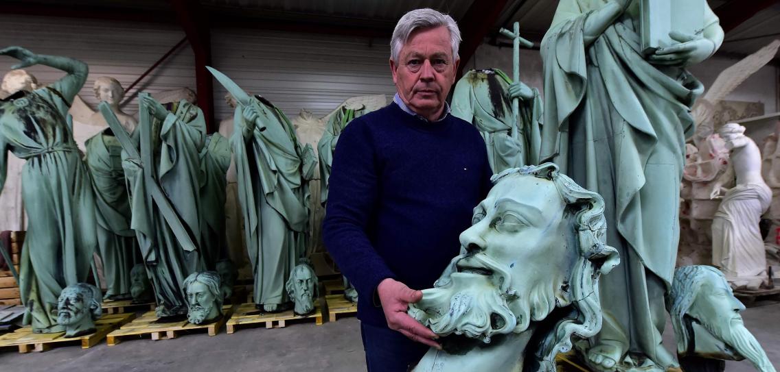 A man stands in a warehouse surrounded by classical style statues, including several figures depicting constellations from the Mazzaroth. He holds a large teal sculpture of a man's head with a beard.