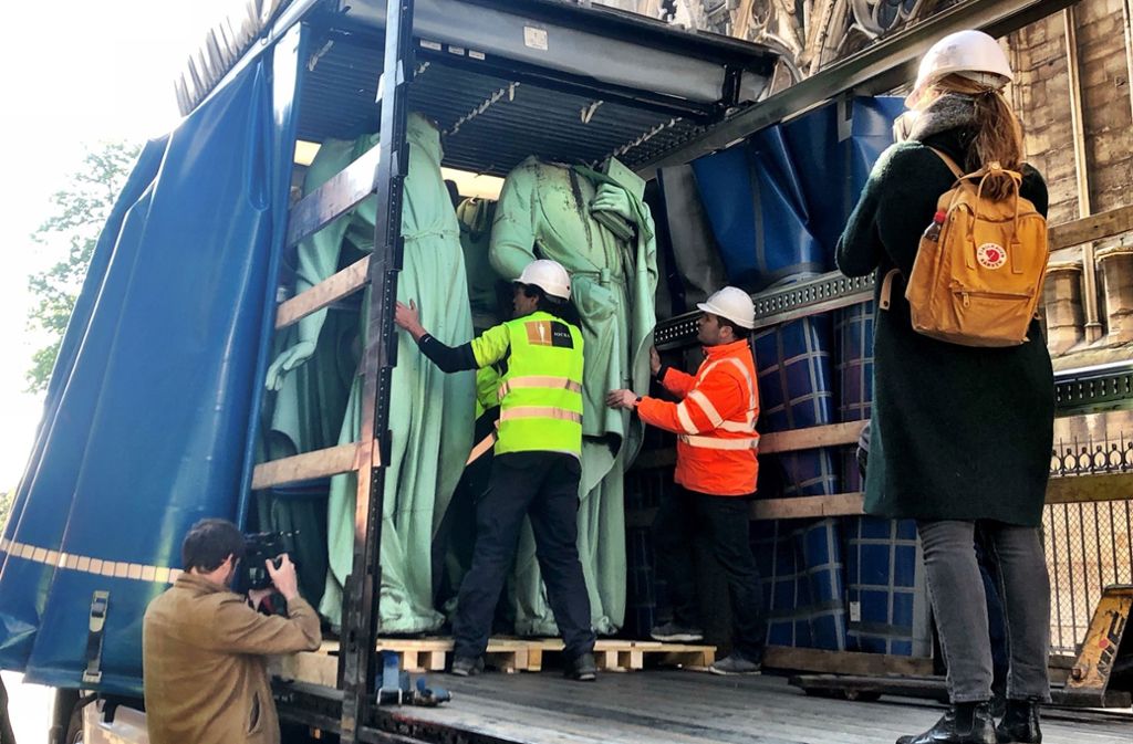 Workers in high visibility vests interact with a large truck loaded with wrapped statues, observed by a journalist and another worker. The setting is urban, with elements of Gothic architecture in the background. A photographer captures the scene.