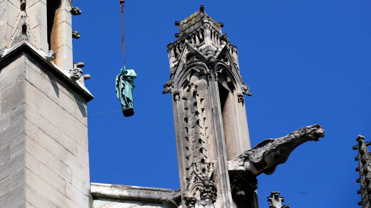 A bronze statue of Virgo, one of the constellations in the Mazzaroth, is suspended by a crane beside a Gothic style building adorned with elaborate stone carvings.