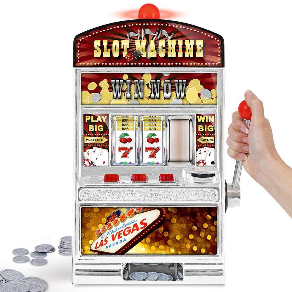A hand operates a miniature slot machine themed with vibrant colors and classic symbols including number sevens and card suits. The top of the machine features a light-up sign saying "Slot Machine Win Now," and a banner below reads "Las Vegas Nevada."