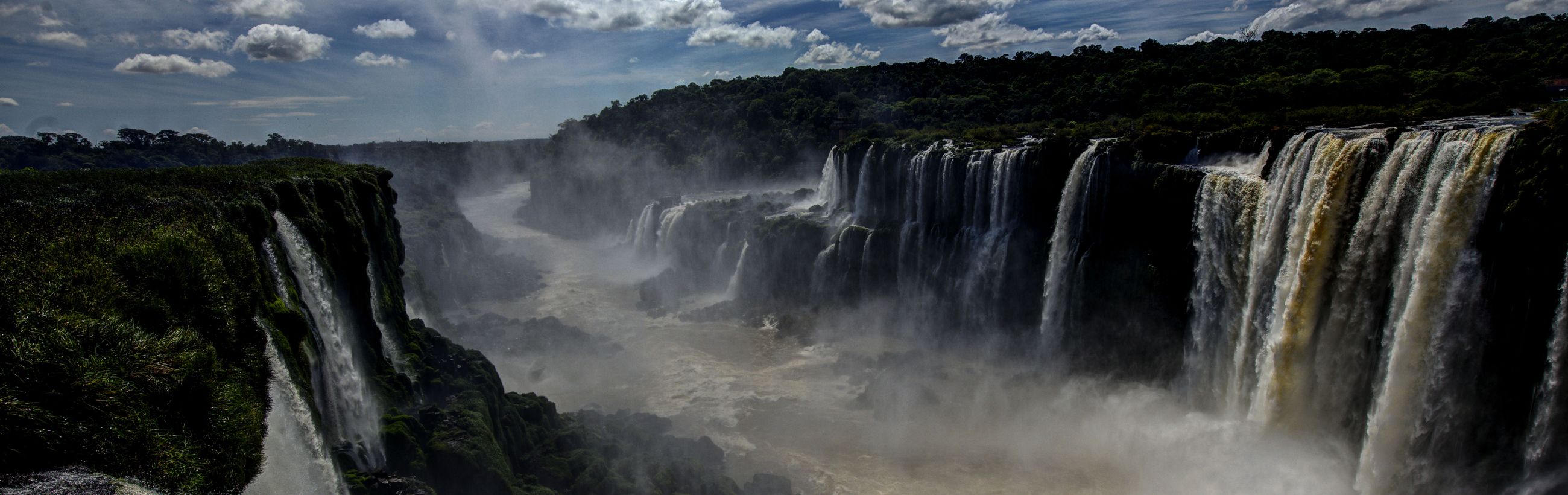 Panoramic view of a majestic waterfall cascading powerfully over a rocky cliff, surrounded by lush greenery under a cloudy sky.
