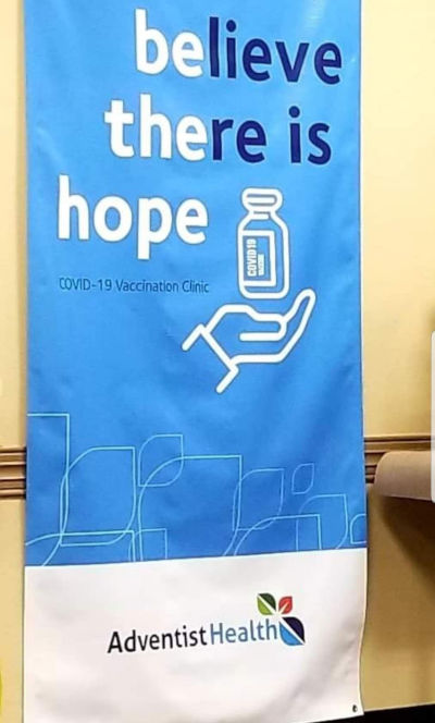 A blue vertical banner stands against a yellow wall, displaying the phrase "believe there is hope" in large white letters at the top, followed by a white graphic of an open hand holding a vial labeled "COVID-19 vaccine". Below, the text reads "COVID-19 Vaccination Clinic" with the logo of Adventist Health at the bottom.
