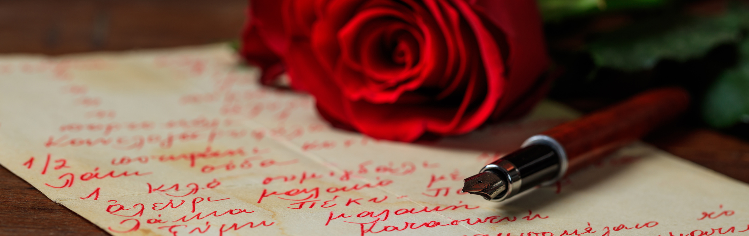 A vibrant red rose lies next to a handwritten letter in red ink on a wooden surface, with a fountain pen positioned at the end of the text, suggesting a pause in writing. The scene captures a moment of thoughtfulness or romance, devoid of any explicit celestial symbols.