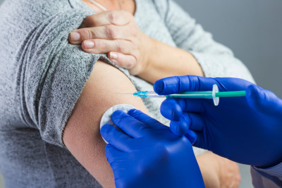 A healthcare professional in blue gloves administering an injection to a patient's upper arm, with the patient gently holding the injection site.
