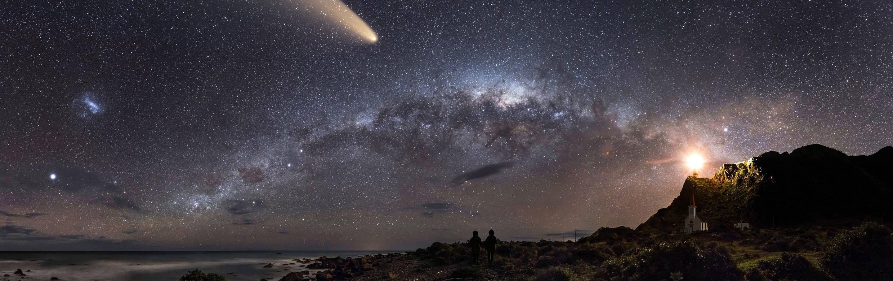 A panoramic night sky featuring a vivid Milky Way galaxy stretches across the scene, accompanied by the bright trail of a comet. Several celestial bodies illuminate the heavens. In the foreground, two silhouetted figures stand observing the spectacle. A lighthouse beams a strong light from a rocky headland, enhancing the dramatic natural and celestial scenery.