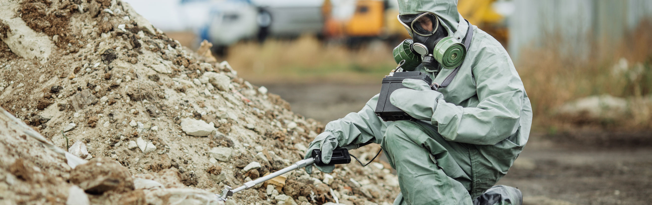 A person in protective hazmat gear crouches at a contaminated site, handling a Geiger counter to measure radiation levels, with piles of debris and a vehicle in the background.