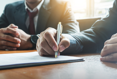Two men seated at a wooden table, one signing a document with a yellow pen while the other observes.