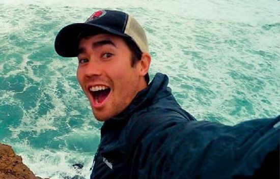 Excited young man in a dark jacket and baseball cap, smiling and extending his arm for a selfie, with a backdrop of spirited ocean waves striking against the shore.