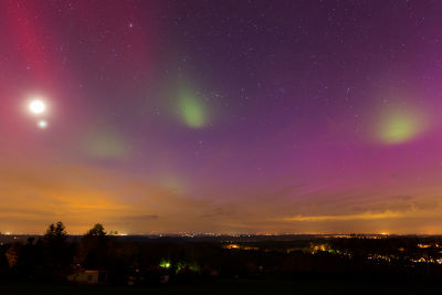Nighttime landscape under a celestial sky featuring bright celestial bodies and multi-colored atmospheric optical phenomena above a horizon of distant city lights and sparse land.