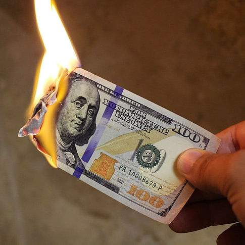 A hand holds a burning 100 US dollar bill, with flames consuming the edge as Benjamin Franklin's portrait is visible.