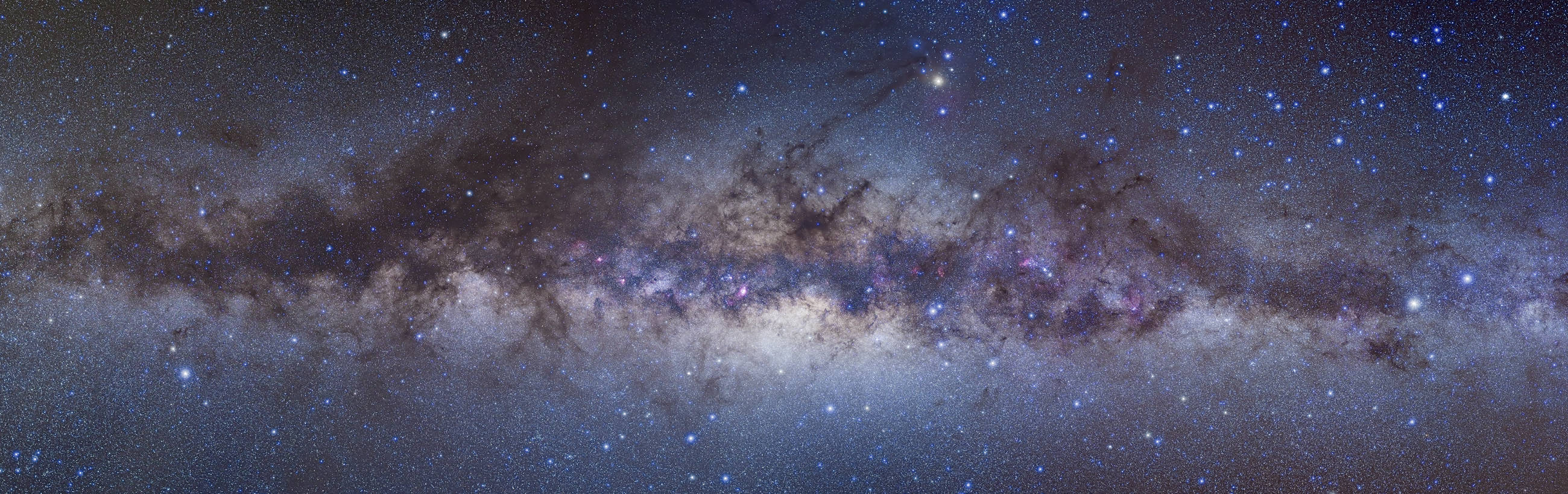 A panoramic view of the Milky Way galaxy showcasing a dense field of stars with clusters of interstellar clouds. The celestial expanse is dotted with myriad stars, nebulae emitting a faint glow, and streaks of cosmic dust.