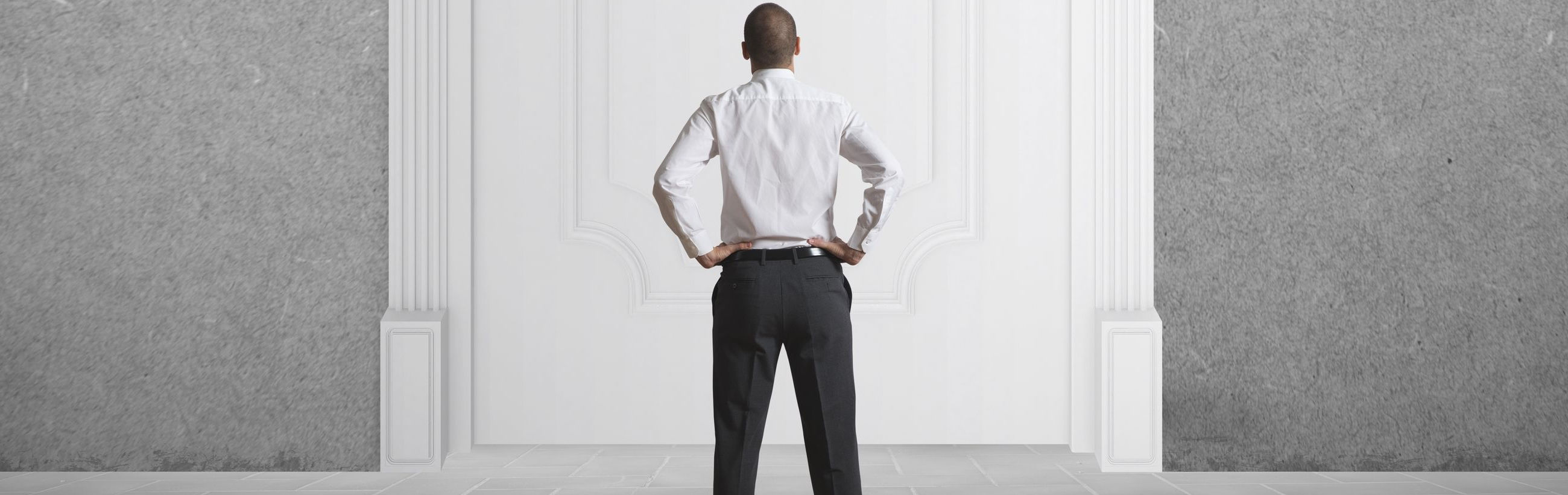 A man in a white shirt and black trousers stands confidently with his hands on his hips, facing towards an ornate white wall with classical architectural details.