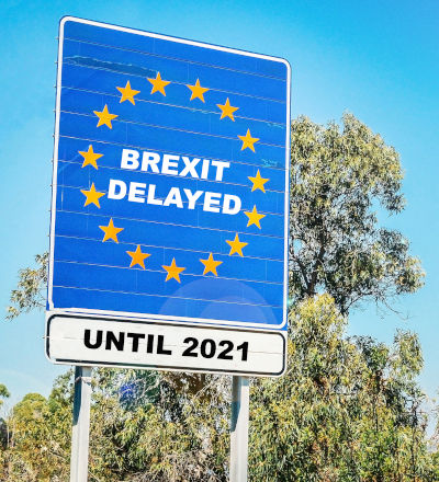 A signboard mimicking the EU flag design with text reading "Brexit Delayed Until 2021" against a backdrop of blue sky and trees.