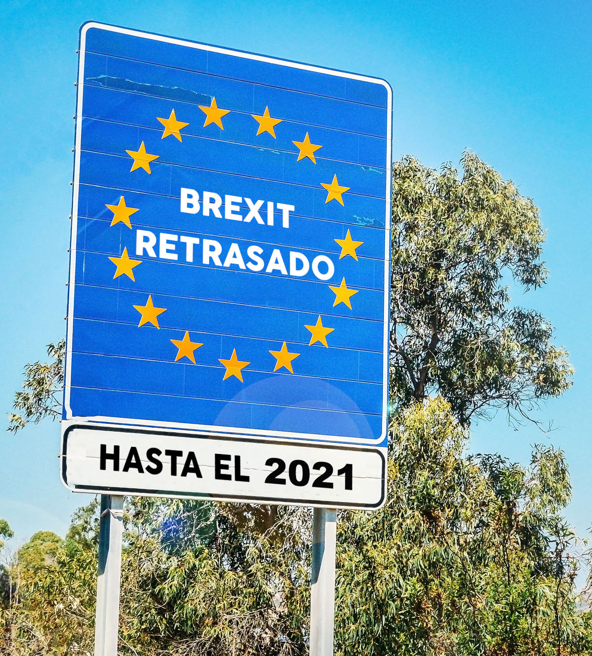 Un cartel grande al aire libre que muestra un mensaje sobre "Brexit Retrasado" con fecha hasta el 2021, decorado con estrellas doradas sobre un fondo azul, ubicado ante un cielo claro y árboles verdes.