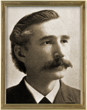 Vintage portrait of a mustachioed man with neatly styled hair, framed in an elegant golden frame. The man is wearing a formal suit and gazes to his right with a thoughtful expression.