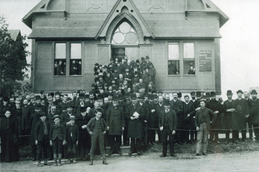 Historical black and white photo showing a large group of people, including adults and children, standing and seated in front of an ornate building with a steep gable and large windows.