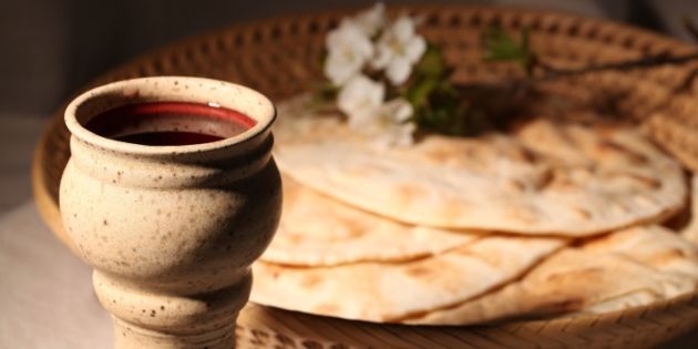 A clay goblet filled with wine beside a woven basket full of unleavened bread and decorated with white blossoms, evoking elements of ancient Biblical feasts.