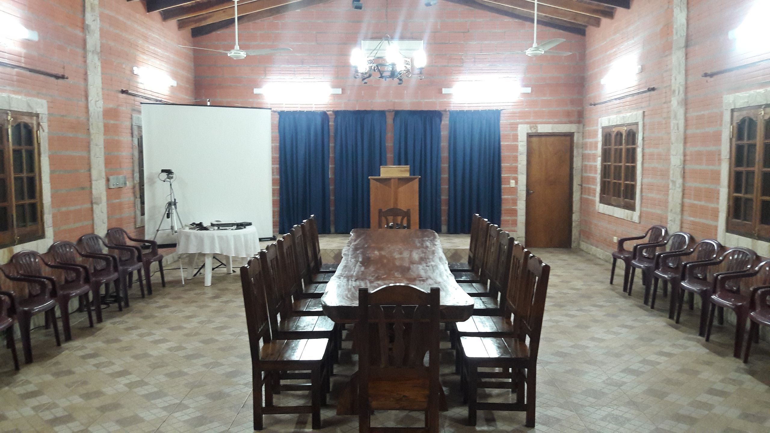 A spacious meeting room featuring exposed brick walls and a high ceiling with wooden beams. A long wooden table flanked by rows of wooden chairs dominates the center, facing a small lectern at the end. Blue curtains cover the windows in the background, and a projector screen along with a camera setup is visible on one side.