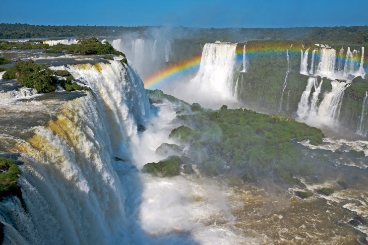A panoramic view of a vast waterfall system with multiple cascades, sending sprays of water high into the sky, where a vibrant rainbow arches across the mist. Dense, lush green vegetation surrounds the waterfalls, under a bright blue sky.