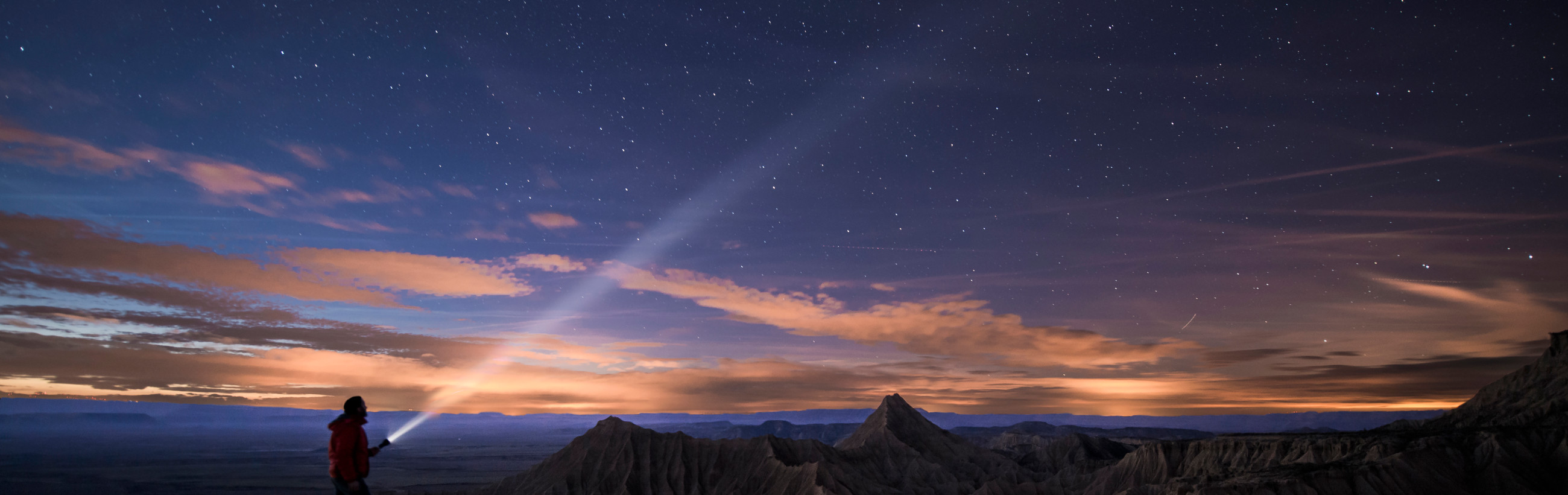 A panoramic view of an expansive landscape at twilight, featuring a clear star-studded sky displaying the Mazzaroth above rugged, eroded mountain ridges. A person in a red jacket is visible in the foreground, gazing towards a luminous, beam-like celestial phenomenon.
