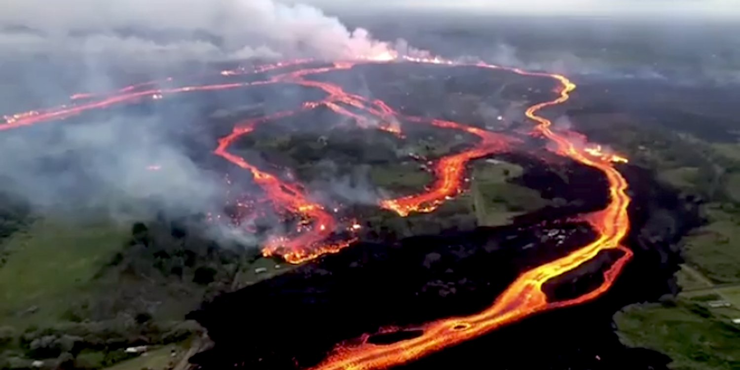 Aerial view of a volcanic eruption showing multiple channels of glowing lava flowing through a landscape, with smoke rising from the active areas.