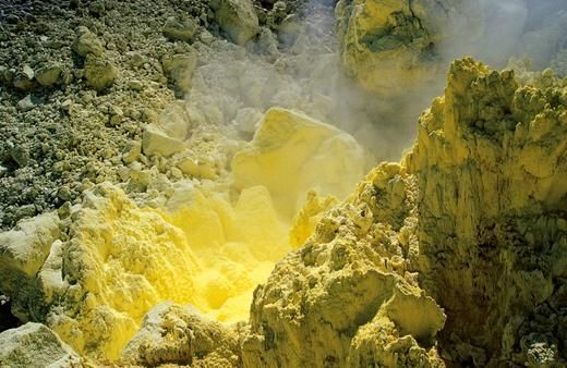 A landscape of a geothermal area featuring vivid yellow sulfur deposits and steam emanating from crevices in rocky terrain.