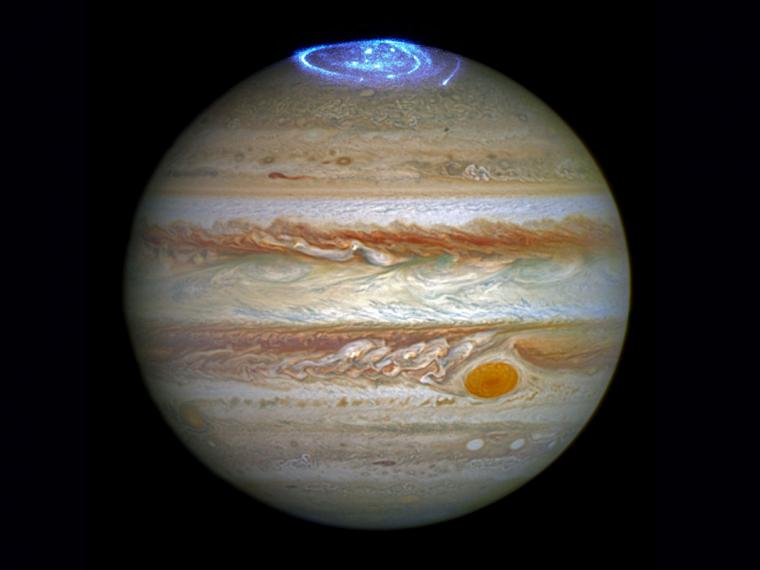 A detailed image of the planet Jupiter, prominently displaying its swirling gas clouds and storms including the famous Great Red Spot. A bright aurora dazzles at the north pole of the planet.