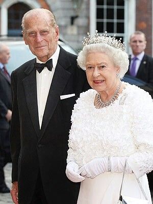 Un hombre mayor con un esmoquin negro está junto a una mujer mayor con un elegante vestido blanco adornado con perlas y encaje, que lleva una delicada tiara y guantes, en un evento formal.