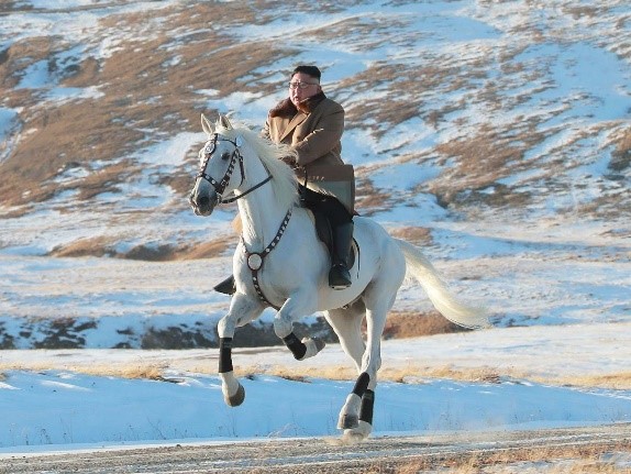 Un hombre con un abrigo beige y guantes negros monta un caballo blanco al galope a través de un paisaje nevado, con colinas cubiertas de hierba marrón al fondo bajo un cielo despejado.