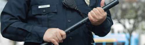 Primer plano de un oficial de policía uniformado que sostiene una porra, con el foco puesto en las manos del oficial y su equipo, incluida una radio sujeta al uniforme.