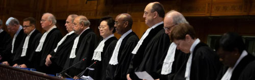 Una fila de nueve funcionarios judiciales, vestidos con túnicas negras tradicionales, inclinando la cabeza respetuosamente en una sala del tribunal revestida con paneles de madera.