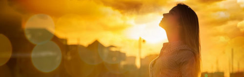 Mujer de perfil frente a un vibrante atardecer, con el rostro vuelto hacia arriba, contemplando. El cielo irradia tonos dorados mientras la luz se filtra entre las nubes, proyectando un cálido resplandor sobre ella y las siluetas de los edificios.