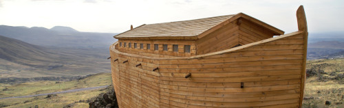 Un arca de madera de gran tamaño, que recuerda al Arca de Noé, situada sobre un terreno rocoso con un telón de fondo de extensas y áridas colinas bajo un cielo nublado.